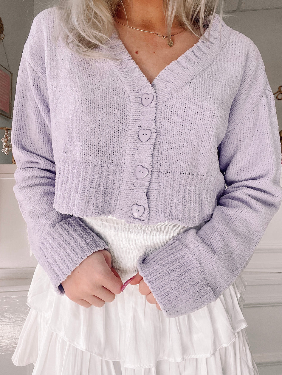 Lavender plush cropped cardigan with heart-shaped buttons, modeled over a white tiered skirt.
