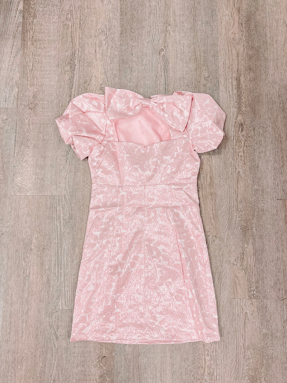 Pink patterned mini dress with puff sleeves and bow detail, laid flat on wood.

