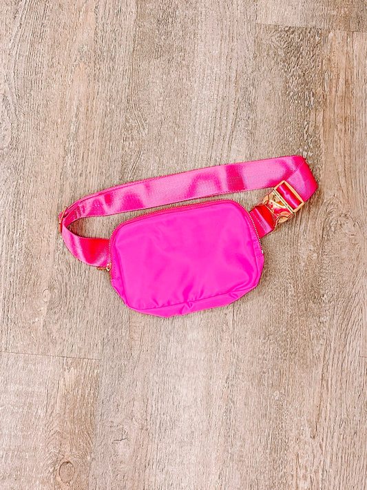 Hot pink nylon belt bag with gold buckle, displayed on wood.
