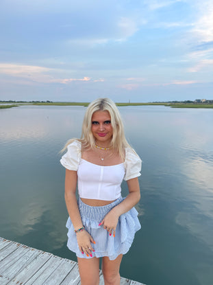 Woman in white puff-sleeve top and blue striped skirt on a wooden dock by the water.
