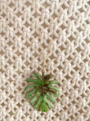 Green monstera leaf pendant necklace on cream knit fabric.
