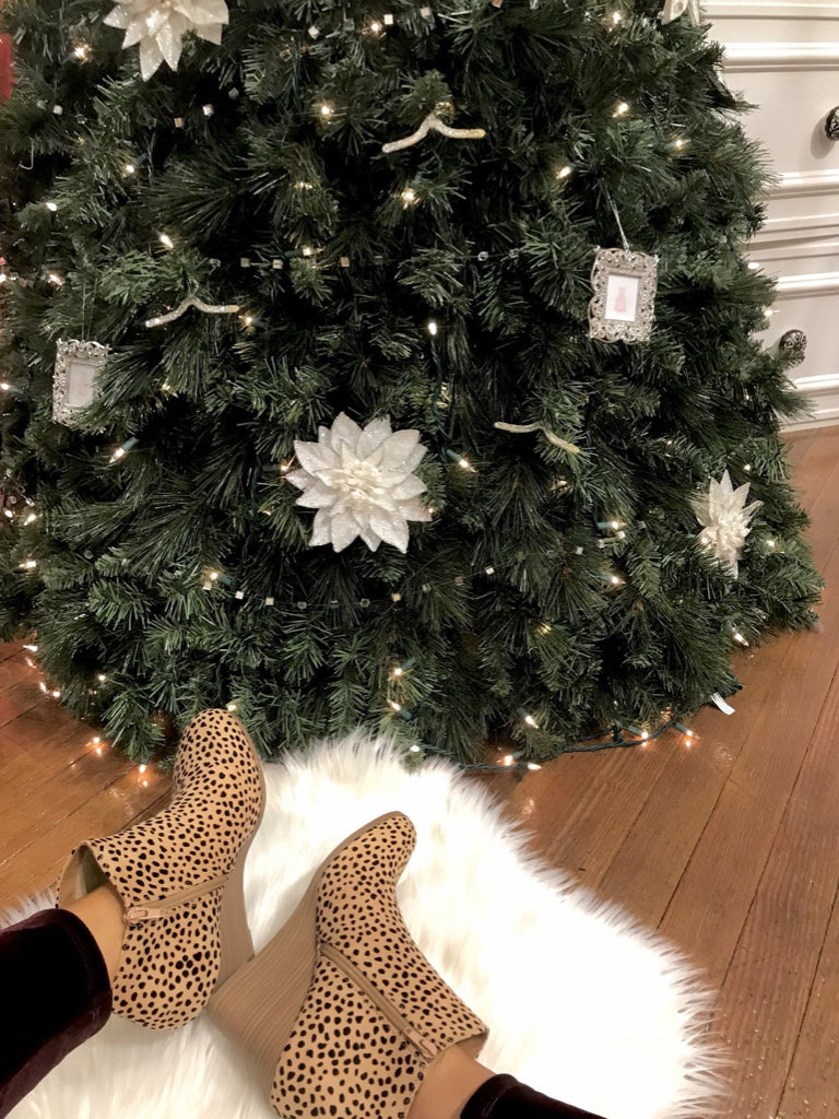 Leopard print booties on a fluffy white rug near a decorated Christmas tree.
