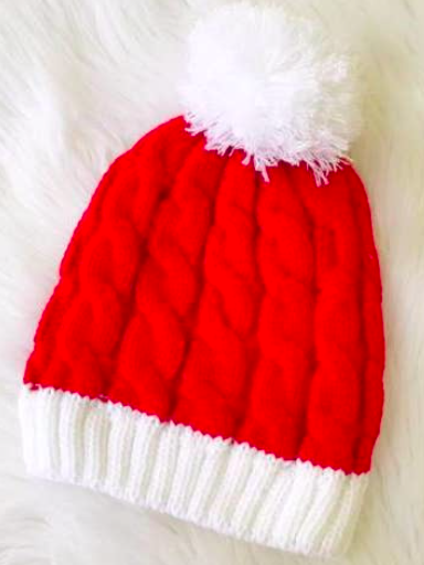 Red cable-knit beanie with white pompom and cuff, on a fluffy white background.

