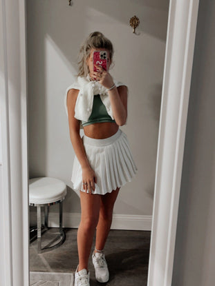White pleated tennis skirt styled with a green top and white sweater, in a dressing room mirror.
