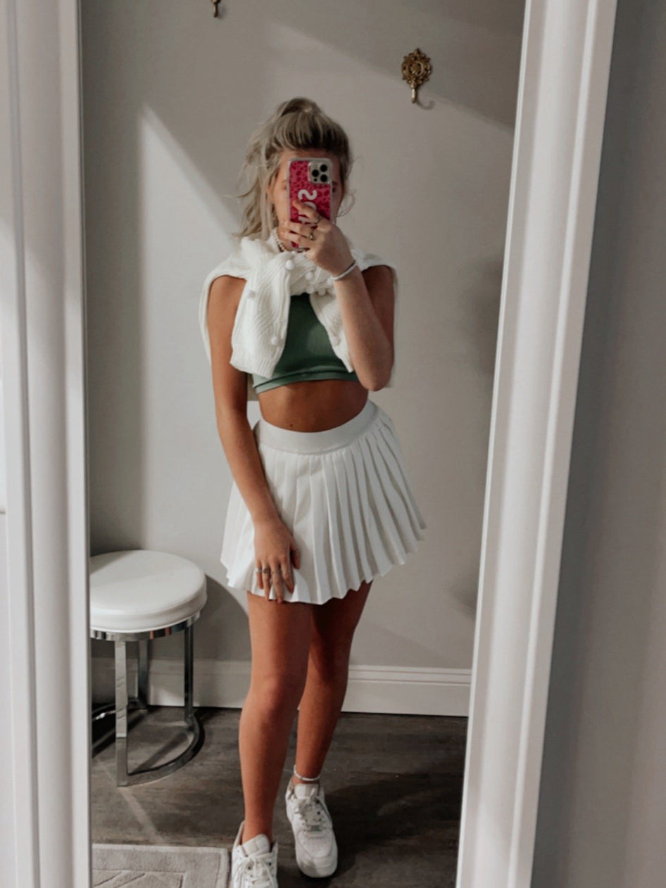 White pleated tennis skirt styled with a green top and white sweater, in a dressing room mirror.
