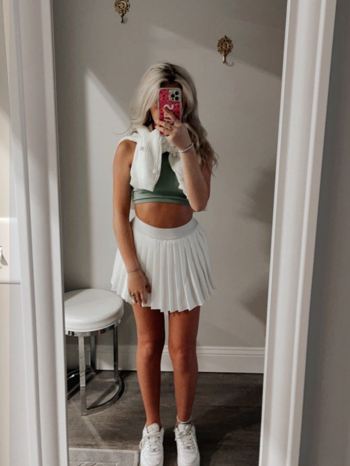 White pleated tennis skirt styled with a sage green top and white cardigan, in a mirror selfie.
