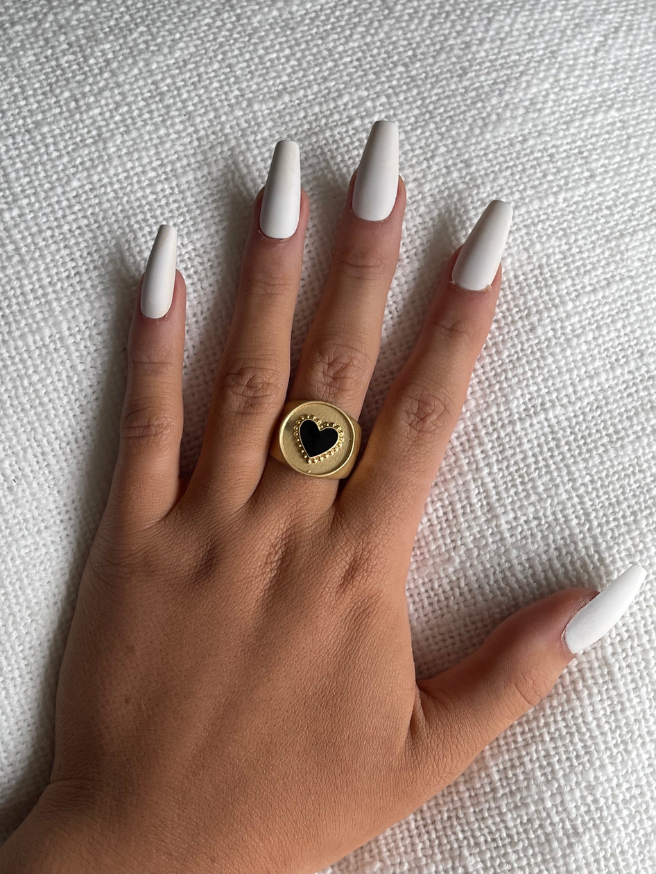 Gold signet ring featuring a black heart, shown on a hand against a white textured background.
