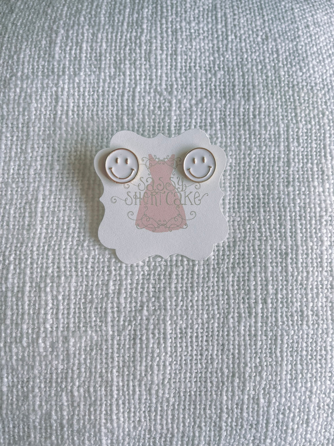 White smiley face stud earrings displayed on a textured white background.
