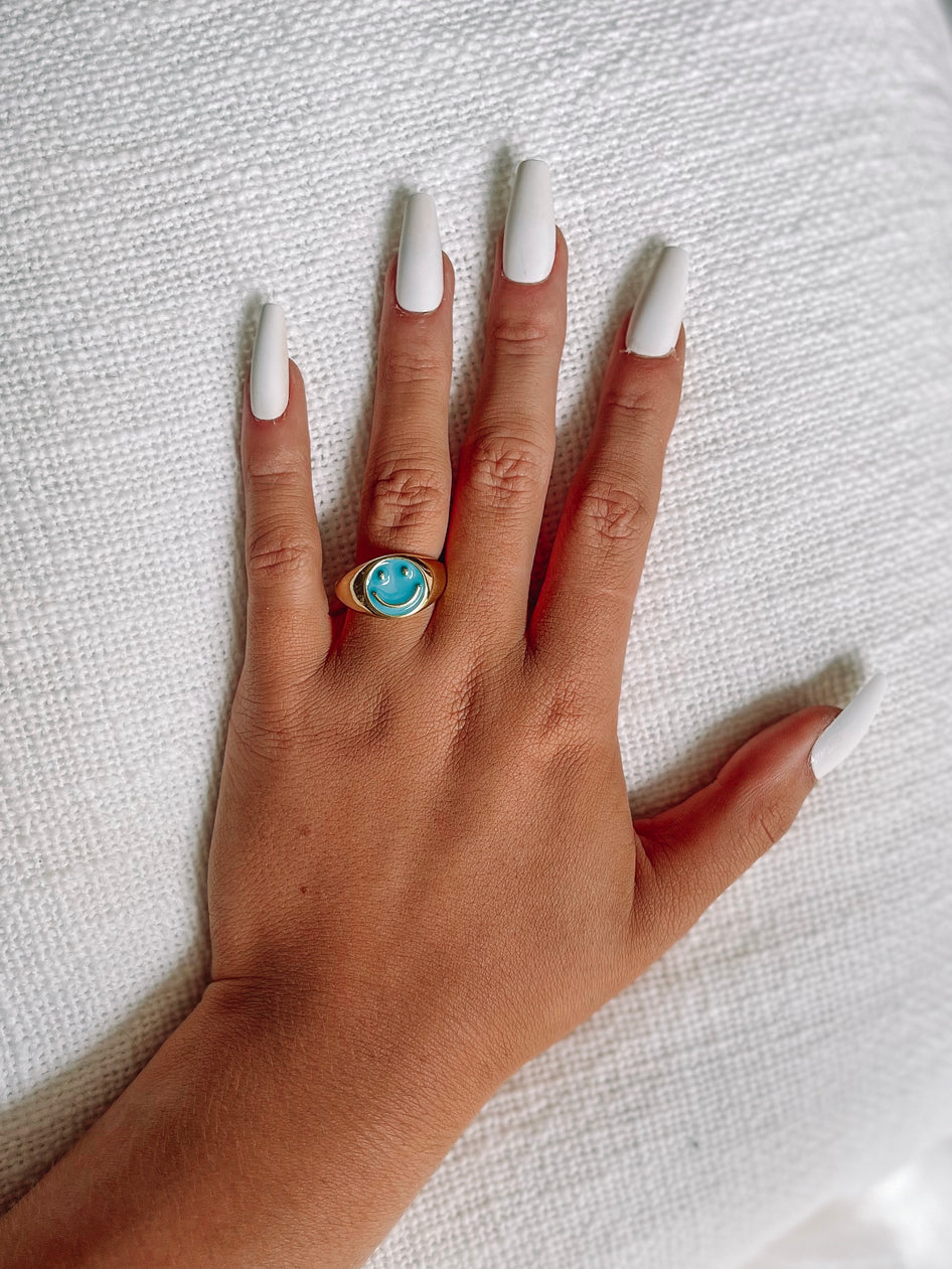 Gold ring with teal smiley face detail, worn on a hand with long white nails, against a white textured background.
