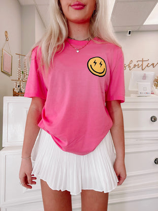 Pink short-sleeve tee featuring a cheerful yellow smiley face graphic, modeled with a white pleated skirt.
