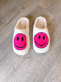 Fluffy white slippers with bright pink smiley faces, shown on a wood floor.

