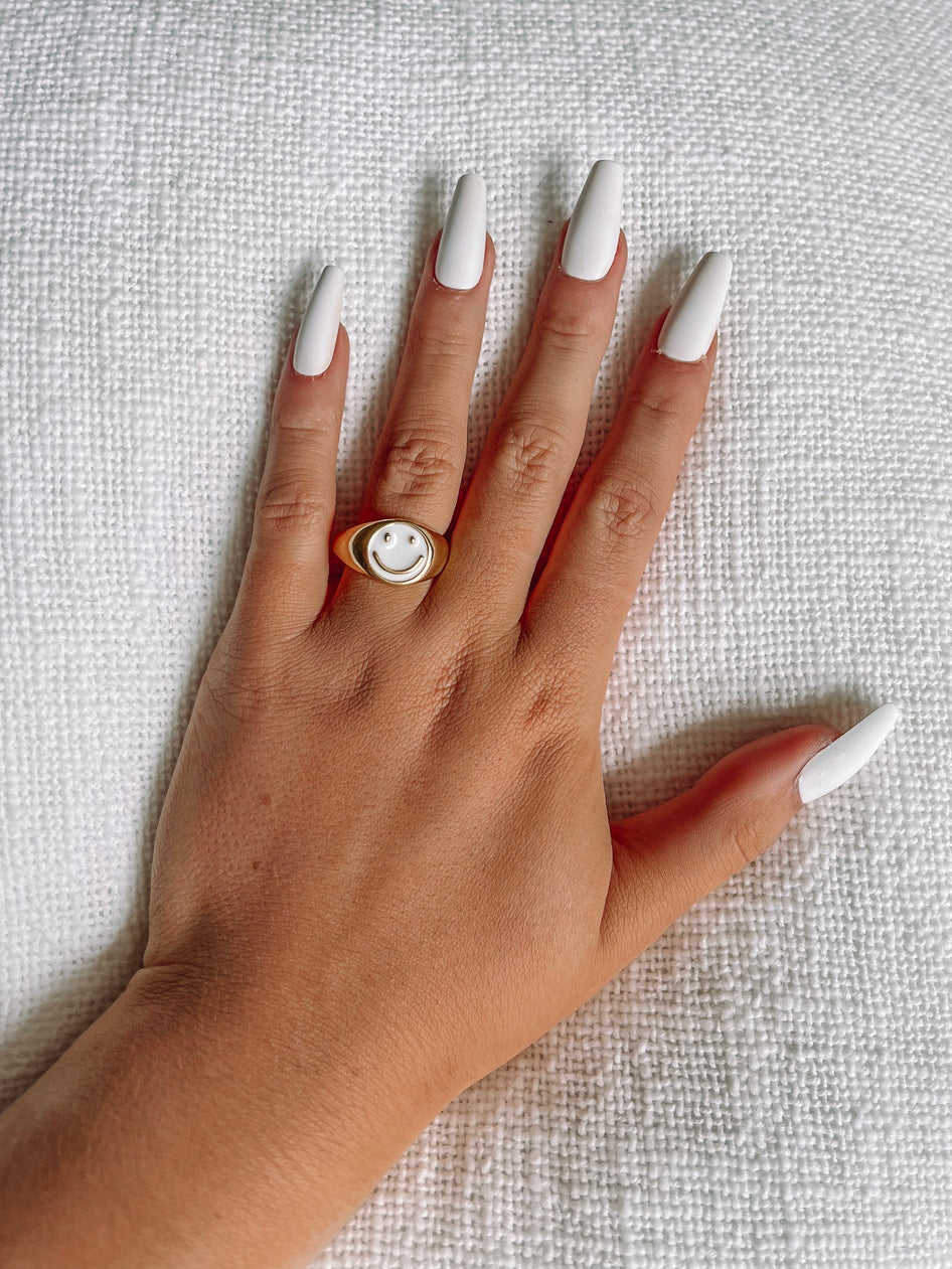 Gold ring with white smiley face accent, displayed on hand with white nails against a white textured background.
