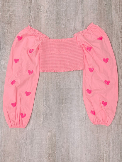 Pink off-the-shoulder top with embellished hearts, long sleeves, and smocked bodice.
