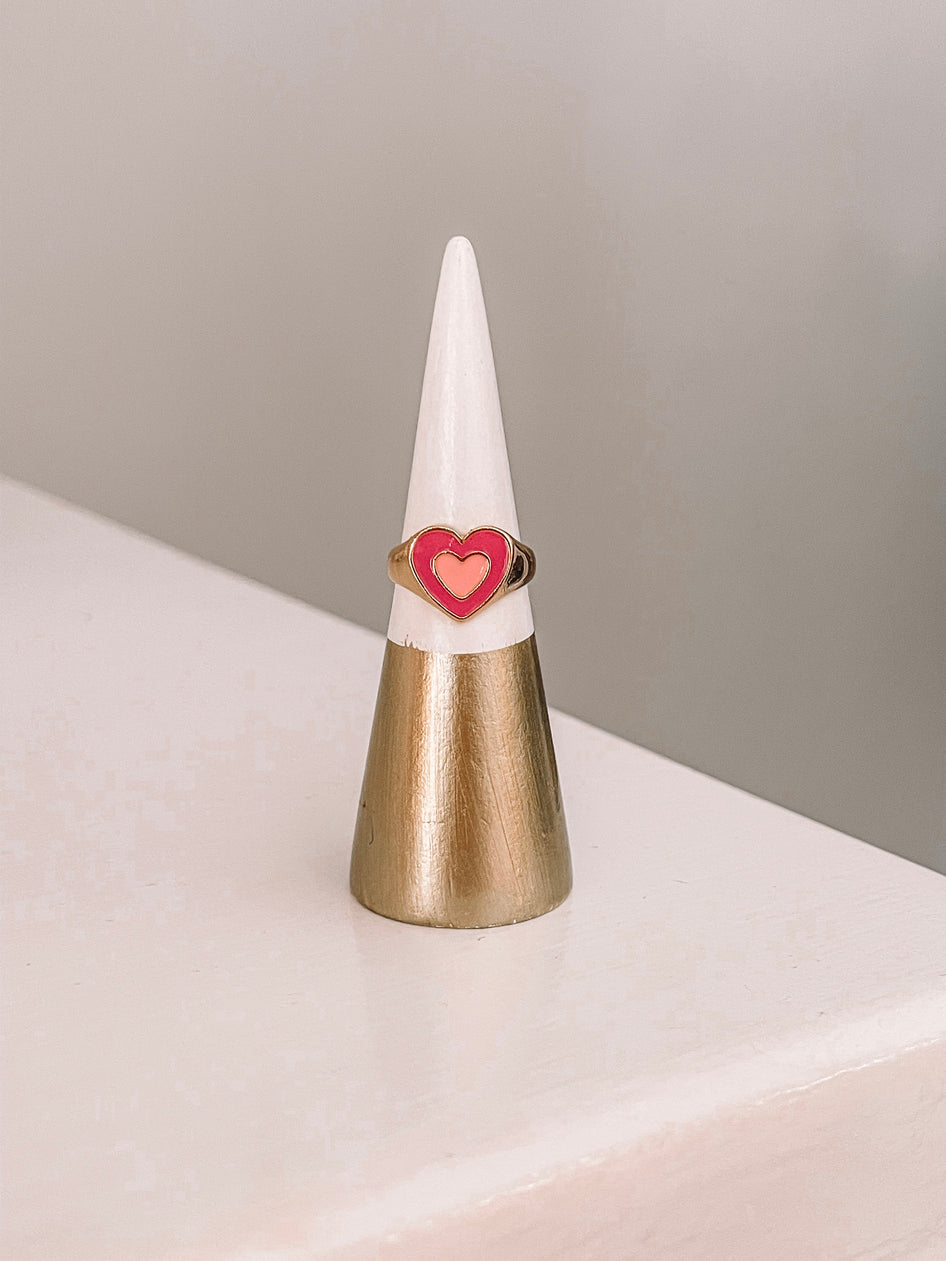 Gold ring with a pink and coral enamel heart, displayed on a ring cone.

