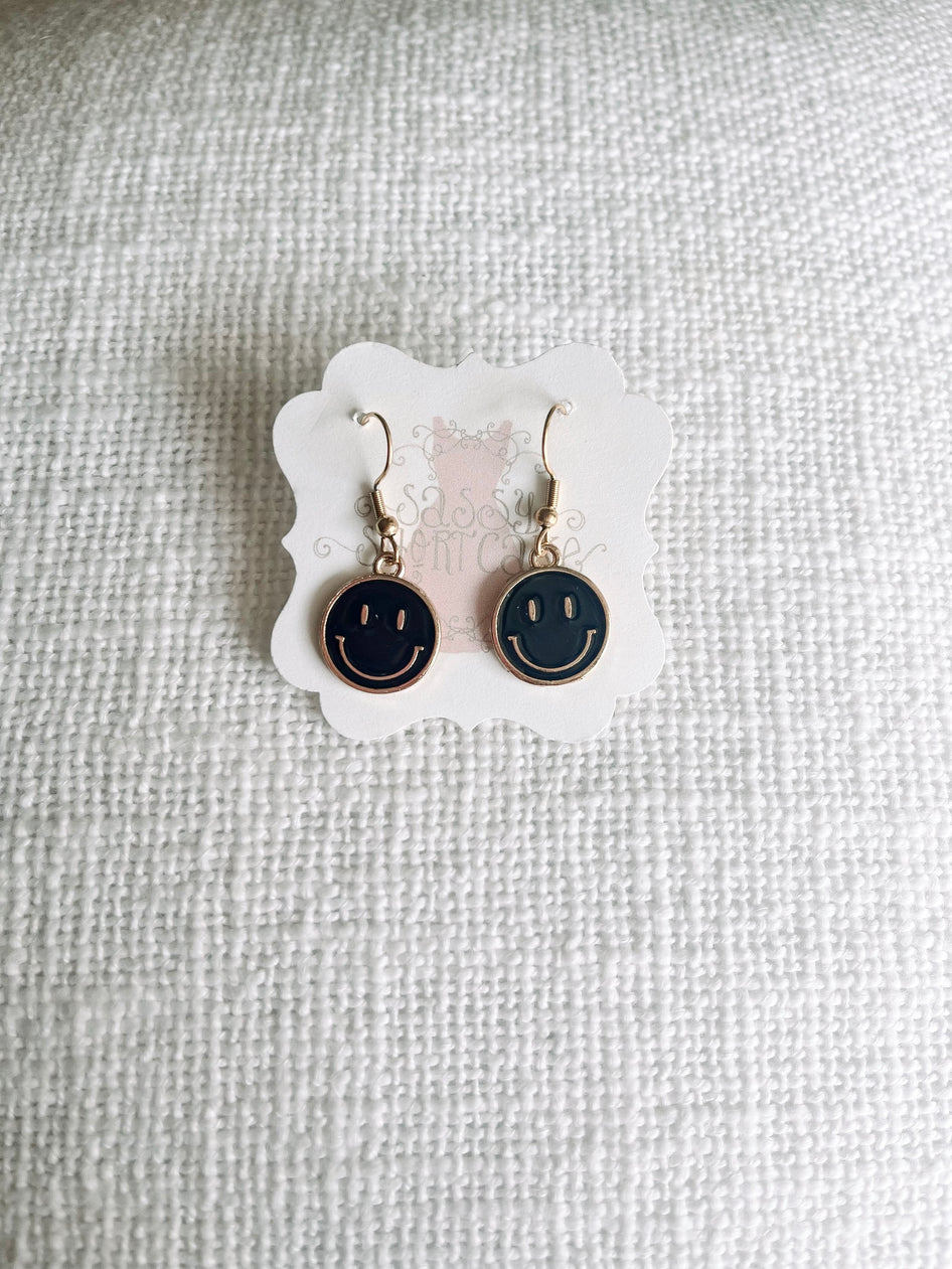 Black smiley face dangle earrings with gold accents, displayed on white fabric.
