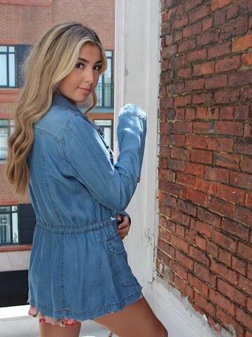 Light wash denim jacket, cinched waist,  model posing near brick wall.
