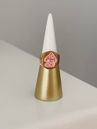 Gold statement ring featuring a pink mushroom design, displayed on a ring cone.
