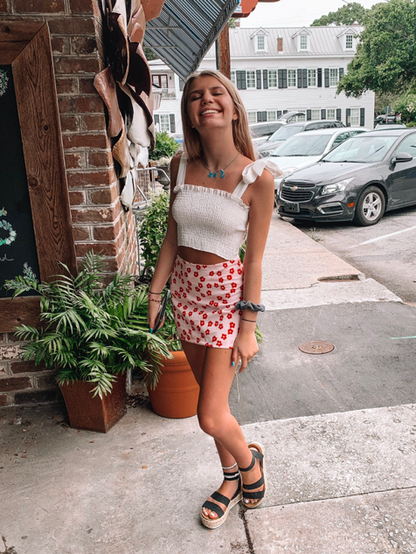 Woman in a white crop top and floral mini skirt, smiling outdoors.
