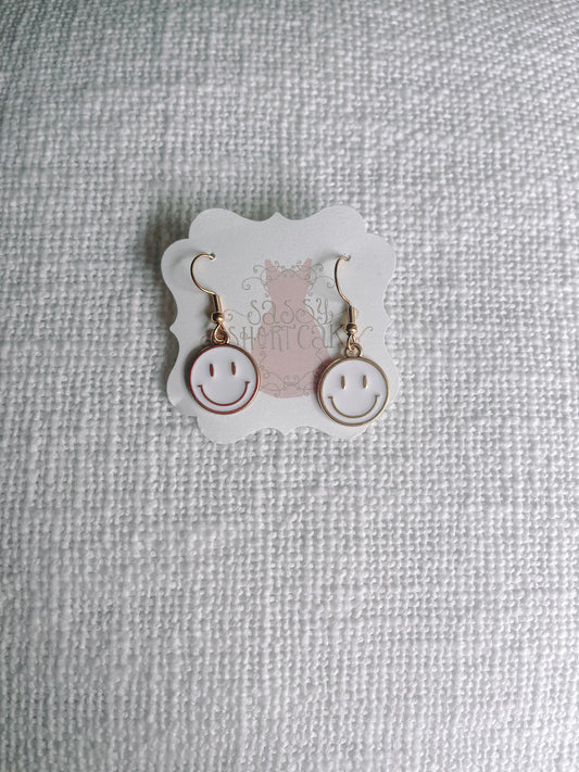 Gold smiley face dangle earrings with white enamel, displayed on textured fabric.