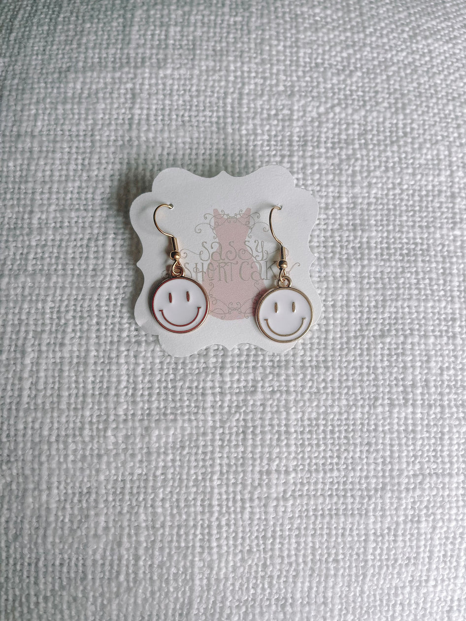 Gold smiley face dangle earrings with white enamel, displayed on textured fabric.
