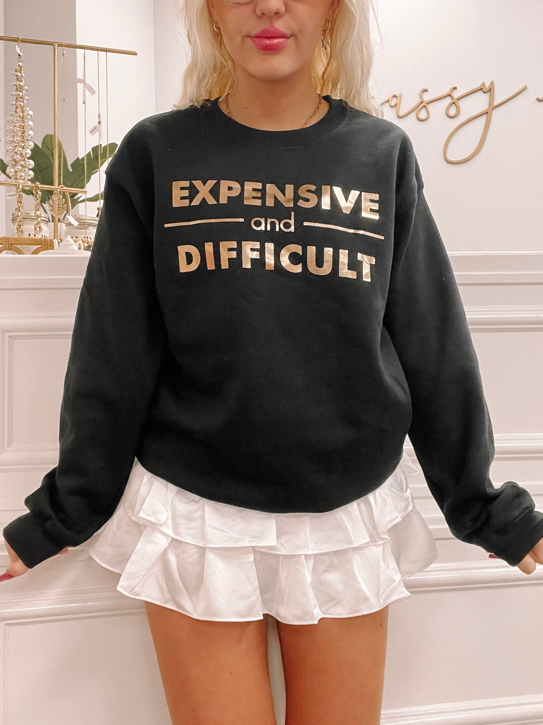 Black crewneck sweatshirt with gold "Expensive and Difficult" lettering, styled with a white pleated skirt.
