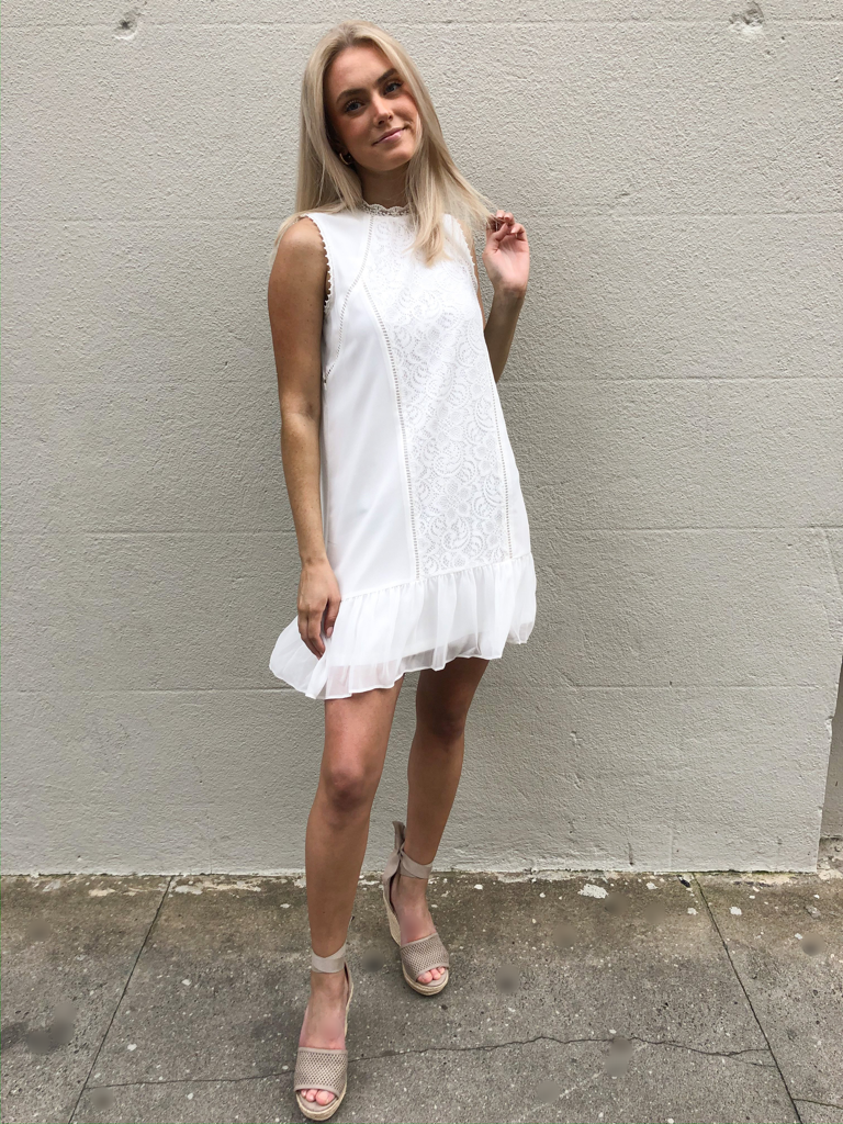 White eyelet dress with ruffle hem, styled with neutral wedges against a textured wall.
