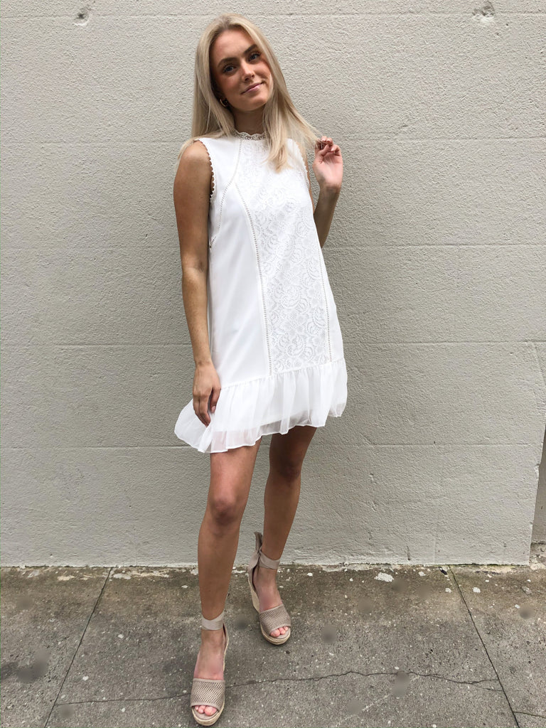 White eyelet dress with ruffle hem, styled with neutral wedges against a textured wall.
