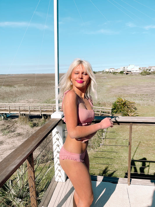 Dusty rose ruffled bikini on a sunny deck overlooking a marshy landscape.