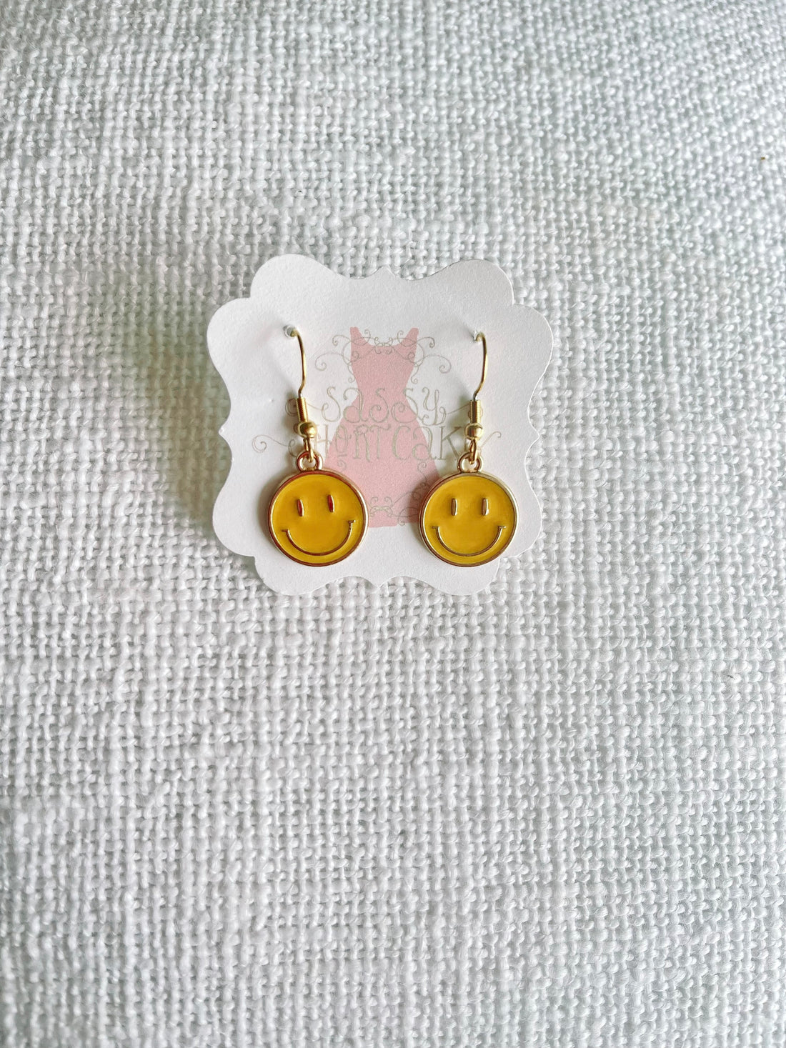 Gold smiley face dangle earrings on a white textured background.
