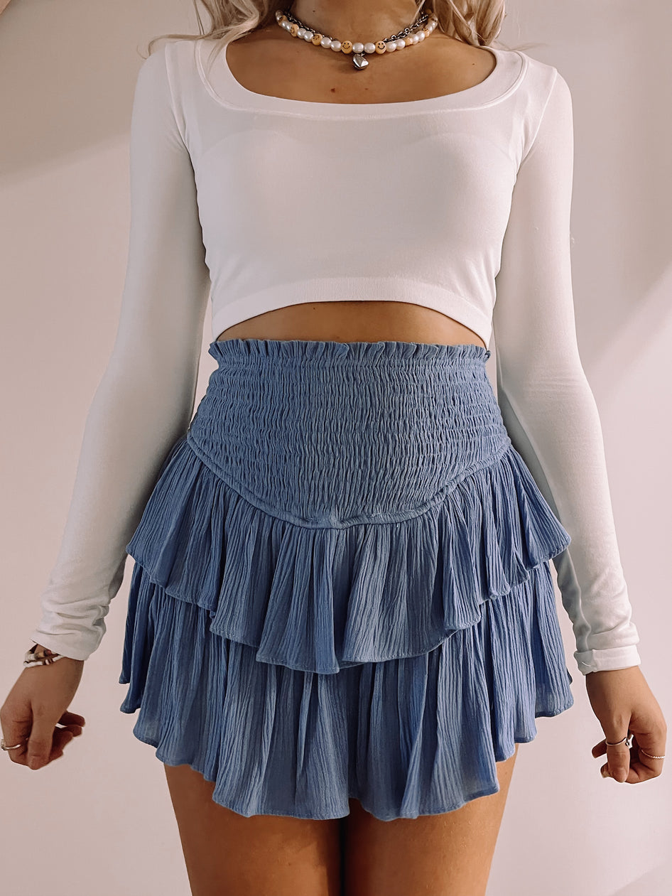 Dusty blue tiered mini skirt with smocked waistband, shown paired with a white long-sleeve top.
