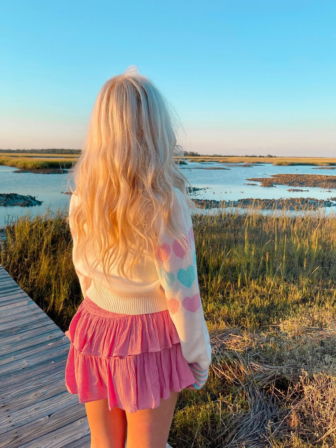 Pastel heart-patterned cream cardigan, worn outdoors by a body of water at sunset.

