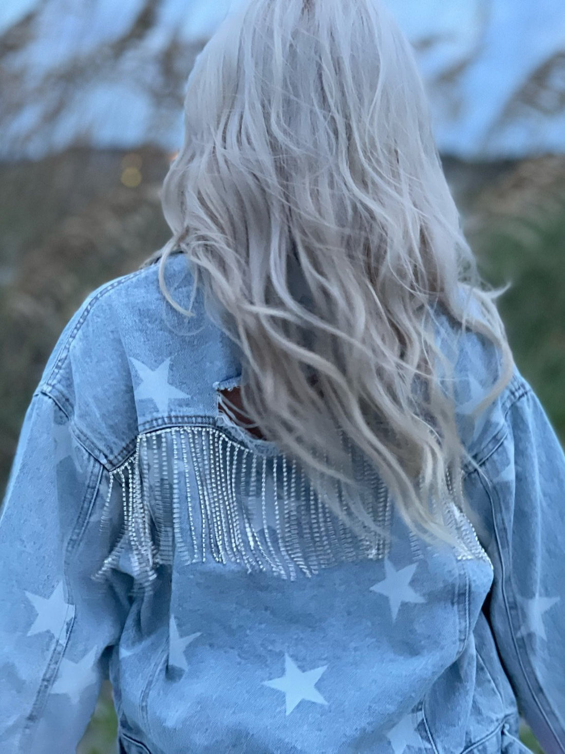 Light wash denim jacket adorned with star print and rhinestone fringe detail, shown from the back.
