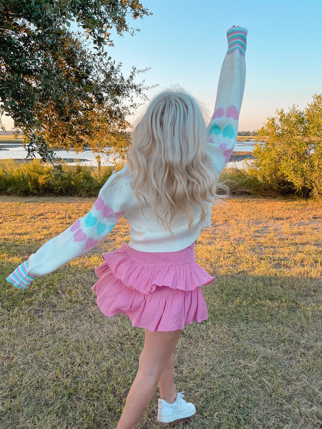 Woman twirling in pastel pink skirt and white sweater with heart details, outdoors near water.
