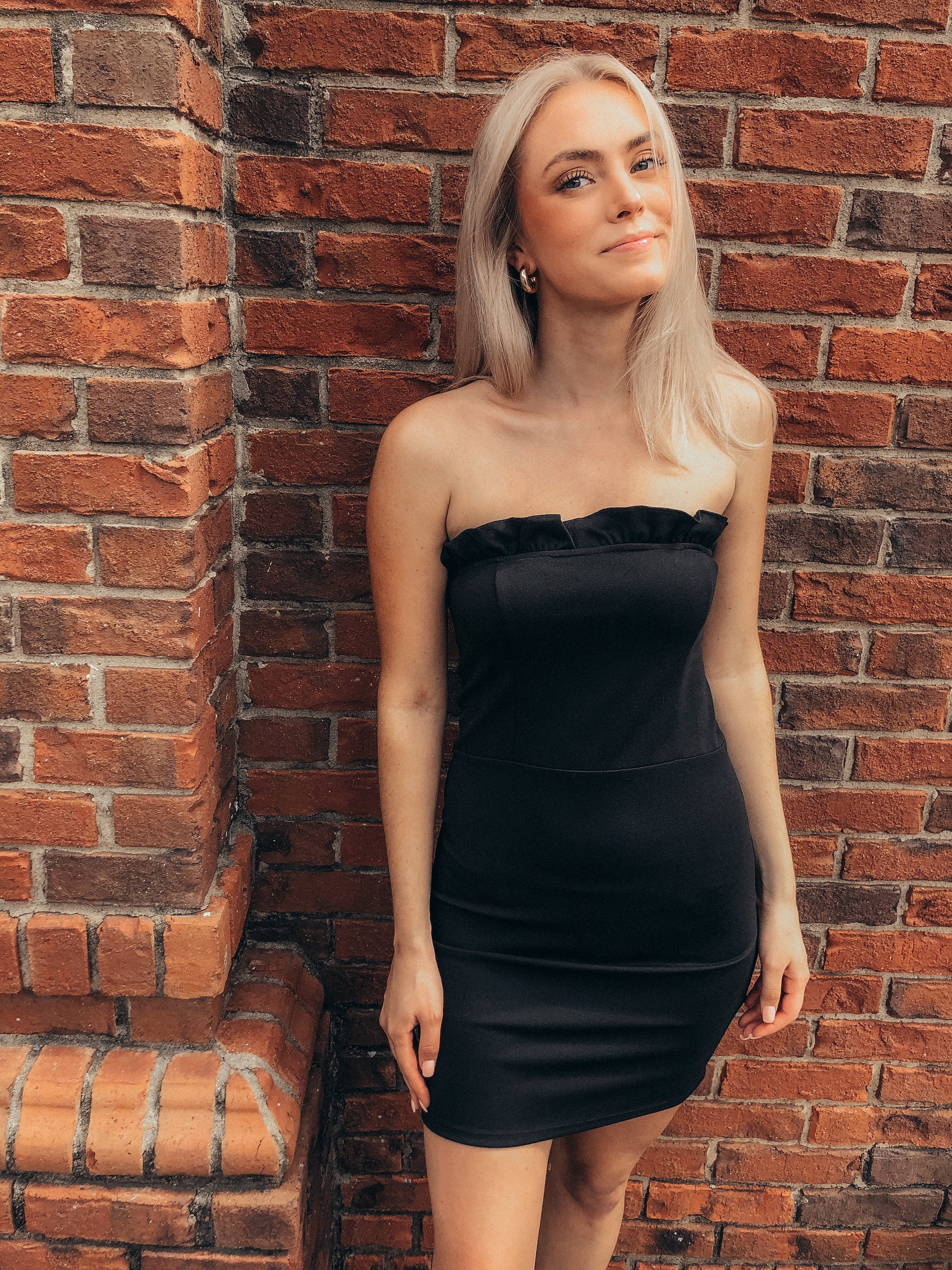 Black mini dress with ruffled neckline, modeled against a brick wall.
