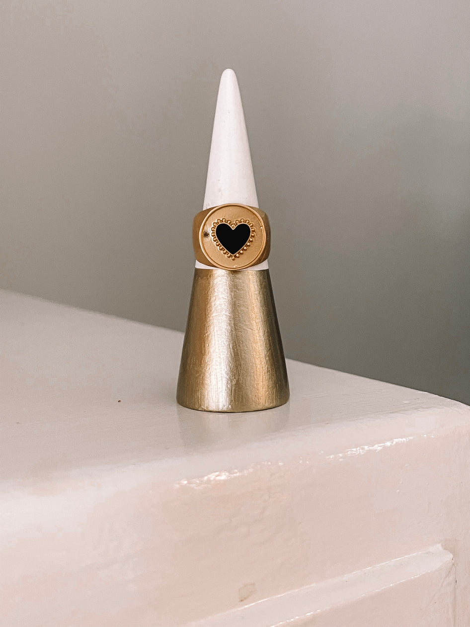 Gold statement ring featuring a black heart, displayed on a gold and white ring cone.

