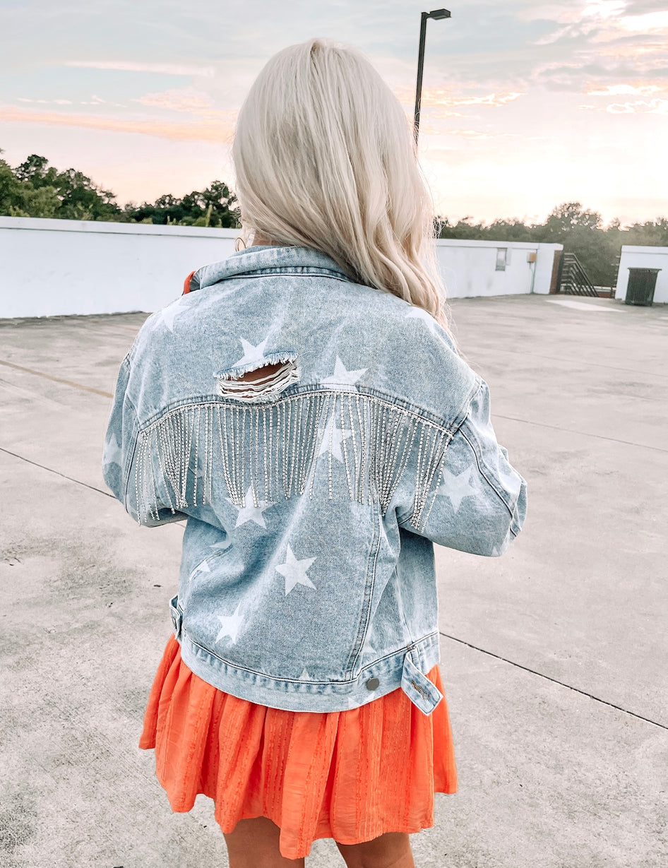 Light wash denim jacket with distressed detail, embellished with rhinestones,  worn outdoors.
