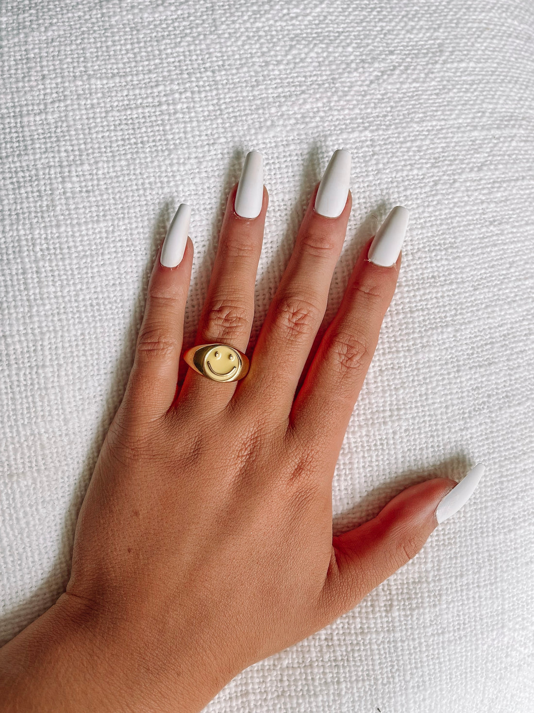 Gold signet ring with a yellow smiley face, shown on a hand against a white textured background.
