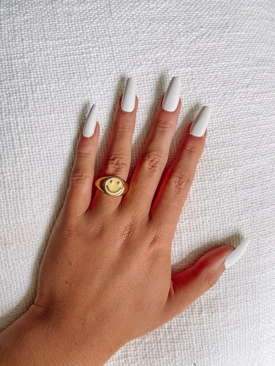 Gold signet ring with a yellow smiley face, shown on a hand against a white textured background.
