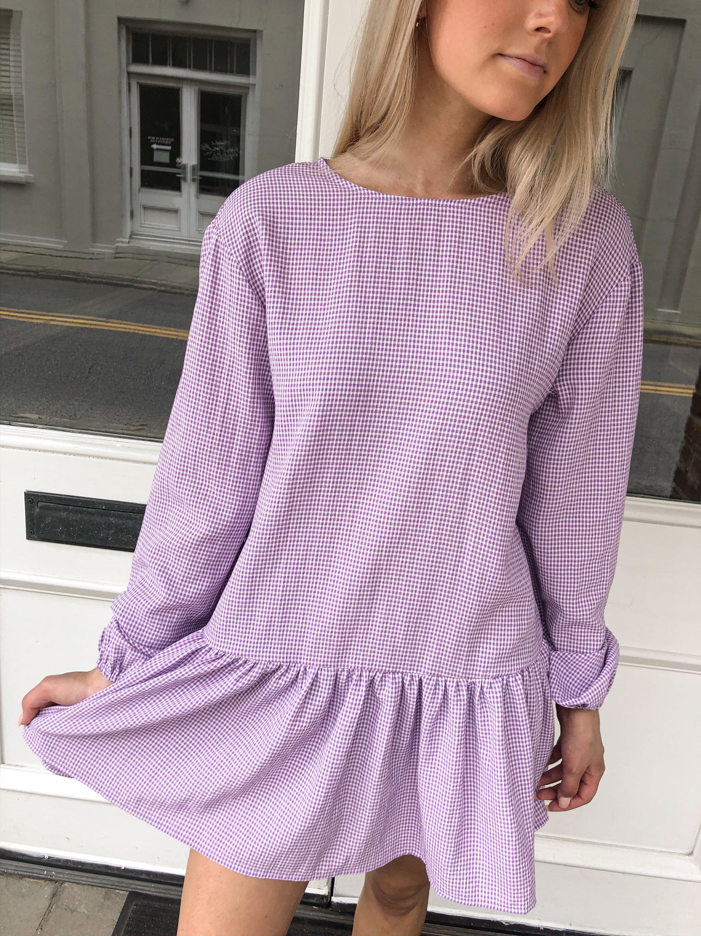 Lavender gingham mini dress with long sleeves and a tiered ruffle hem, shown outdoors.
