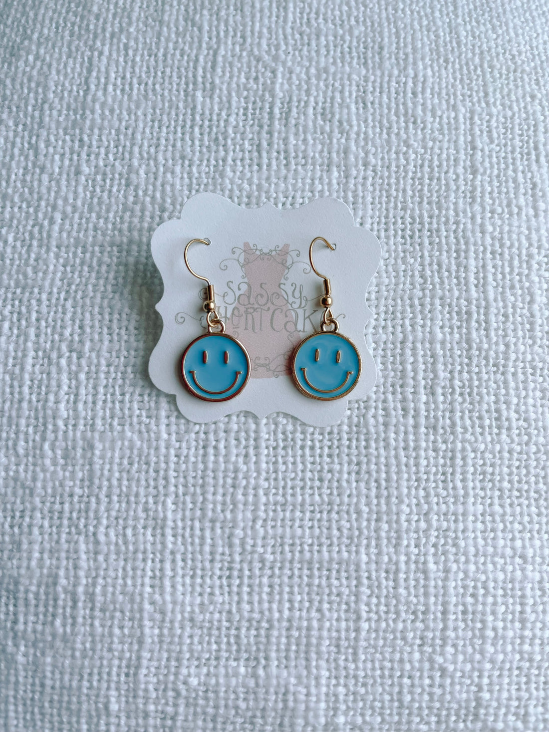 Gold smiley face earrings with light blue enamel, displayed on a textured white background.
