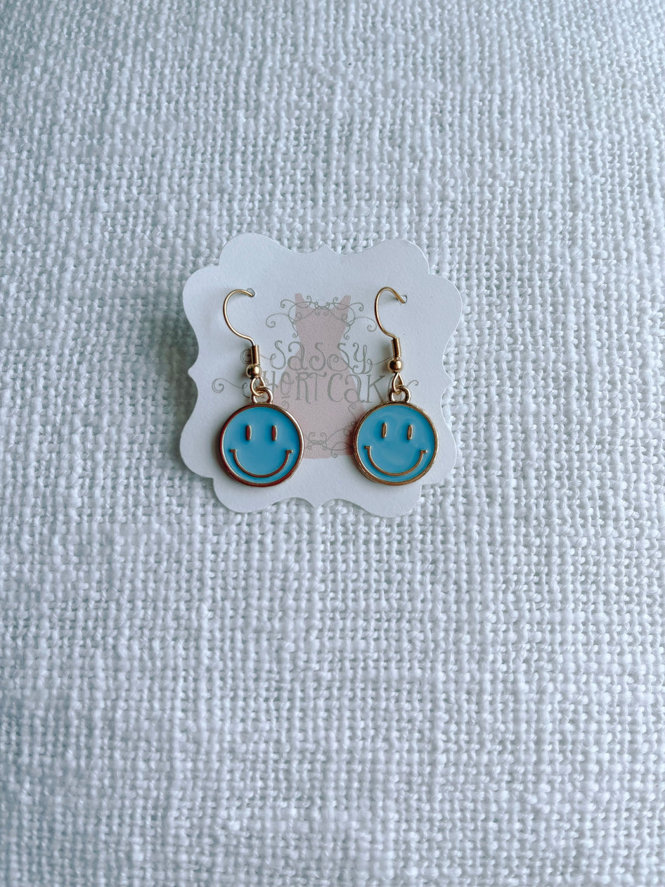Gold smiley face earrings with light blue enamel, displayed on a textured white background.
