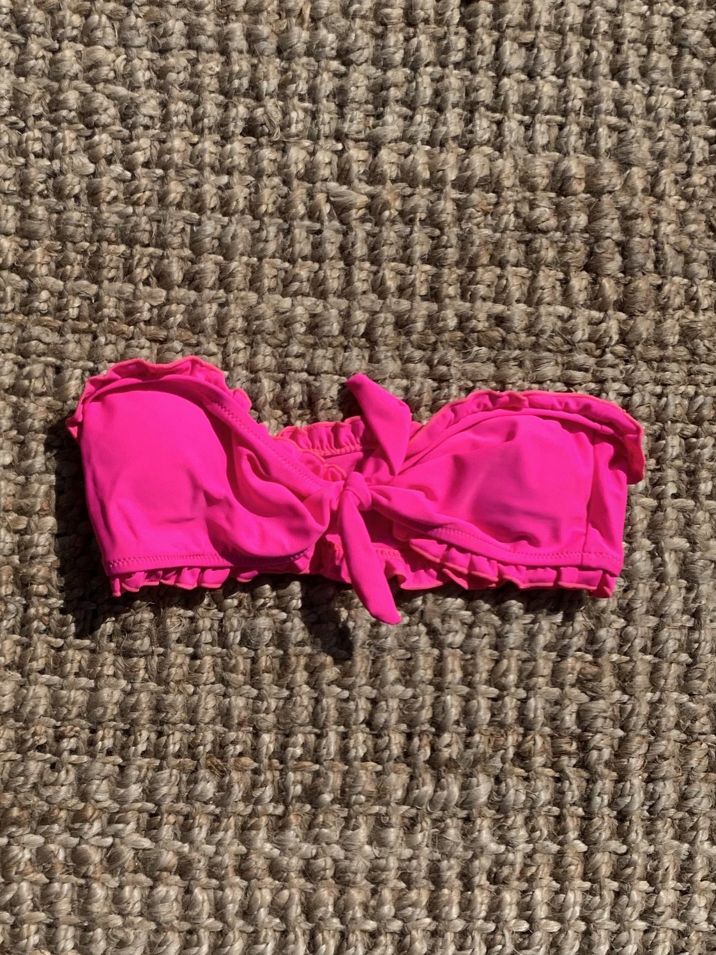 Hot pink ruffle-trimmed bandeau swimsuit top, tied in a bow, shown on a woven surface.
