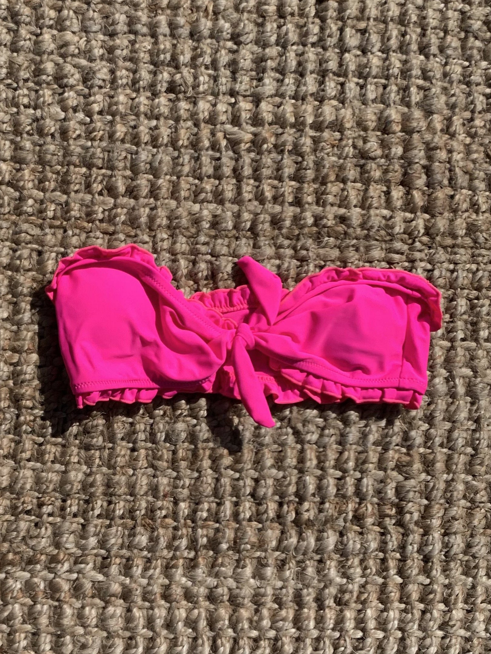 Hot pink ruffle-trimmed bandeau swimsuit top, tied in a bow, shown on a woven surface.
