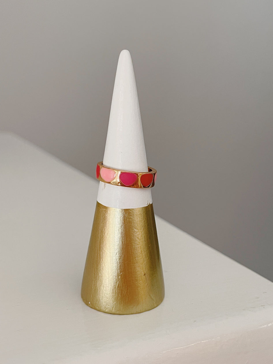 Gold band ring adorned with pink and red enamel hearts, displayed on a gold and white cone.
