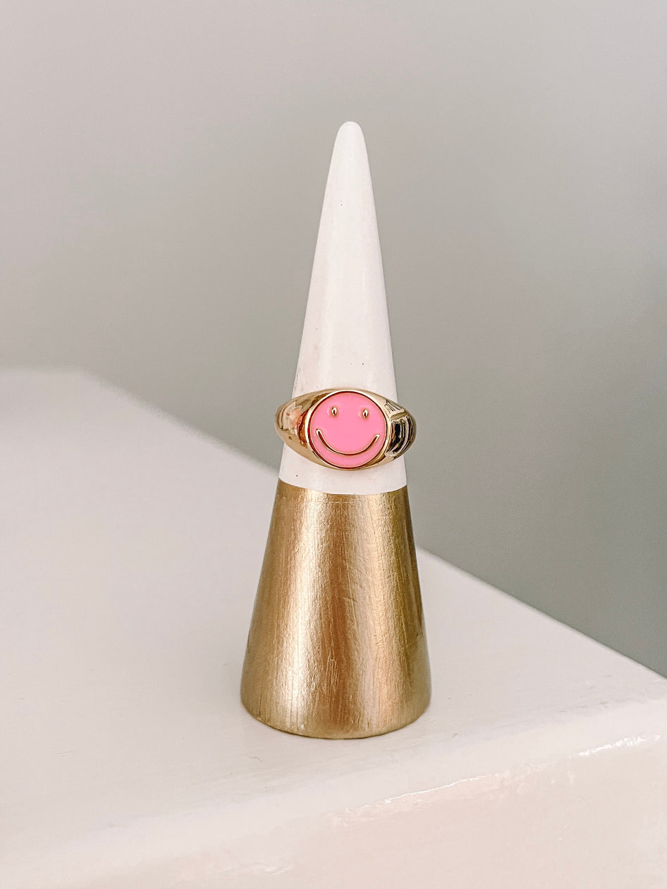 Pink smiley face signet ring displayed on a gold and white ring cone.
