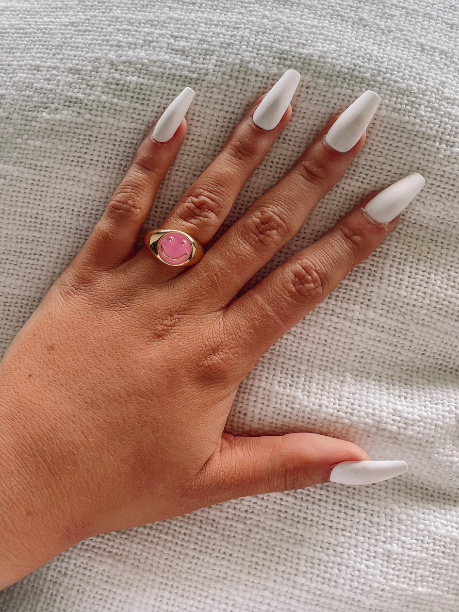 Gold signet ring with pink smiley face, displayed on hand against white fabric.
