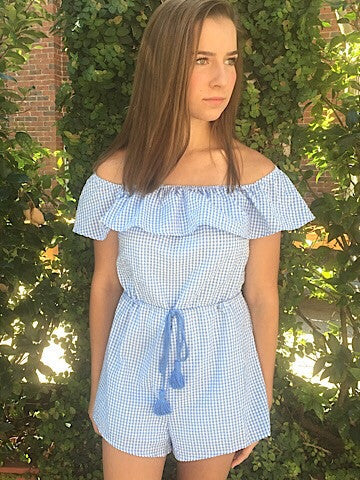 Blue and white gingham romper with off-the-shoulder ruffle and tassel tie waist, modeled outdoors.
