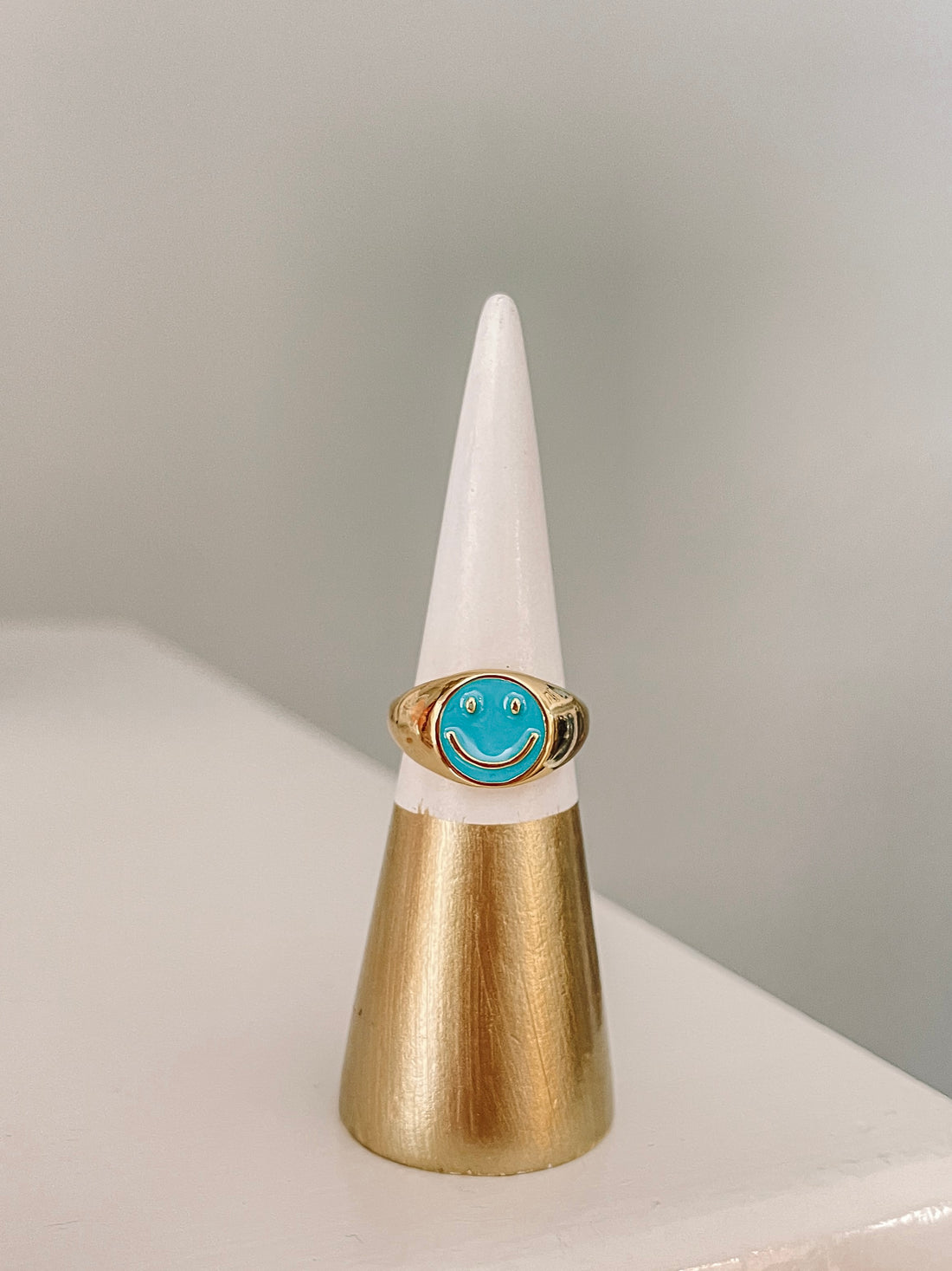 Gold signet ring featuring a turquoise smiley face, displayed on a gold and white ring cone.
