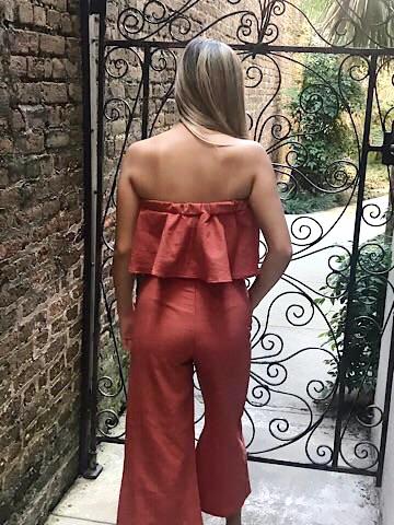 Rust-colored linen jumpsuit with a ruffled strapless top, shown from the back against an ornate iron gate.
