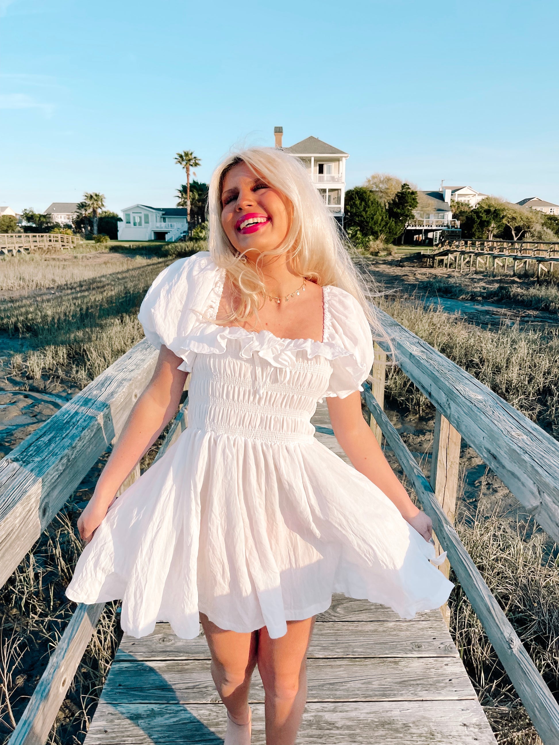White mini dress with puff sleeves and smocked bodice, worn outdoors near water.
